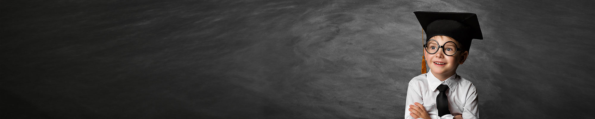 Un jeune enfant souriant porte une toge et une toque de diplômé noires avec un pompon doré. Il a des lunettes rondes, une chemise blanche et une cravate noire, les bras croisés devant un fond gris façon tableau d’école.