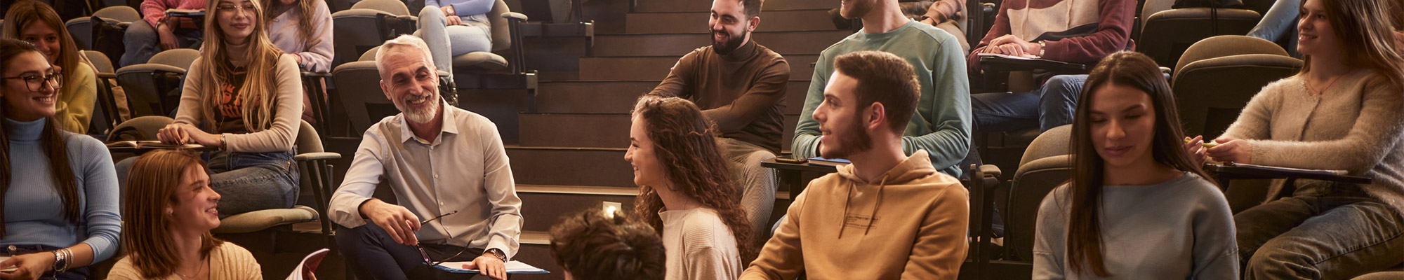 Un professeur d’université assis parmi des étudiants dans un amphithéâtre, échangeant avec eux lors d’une discussion en cours.