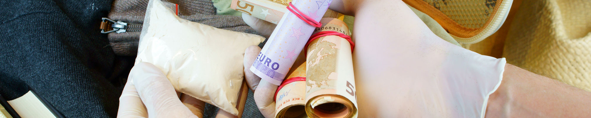 Mains gantées tenant des rouleaux de billets en euros et un sachet suspect dans une valise remplie de vêtements et d’effets personnels